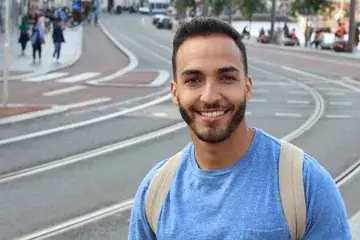A smiling young Hispanic man with a beard wearing a blue t-shirt and backpack, showing a bright healthy smile in a city street setting for a DentaBiome customer testimonial.